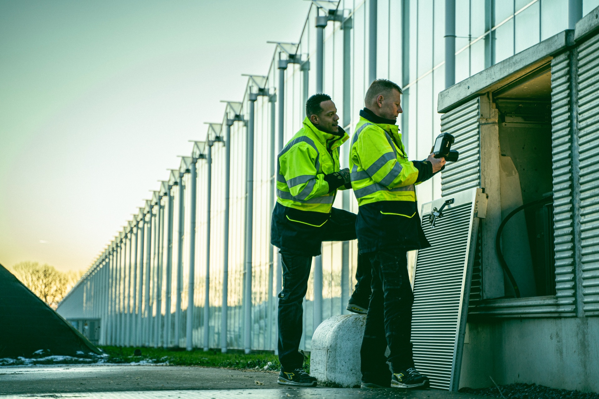 Twee mannen bij een aantal kassen staan voor een compactstation dat open is gemaakt. Via een apparaat kijken zij naar de binnenkant van het compactstation.