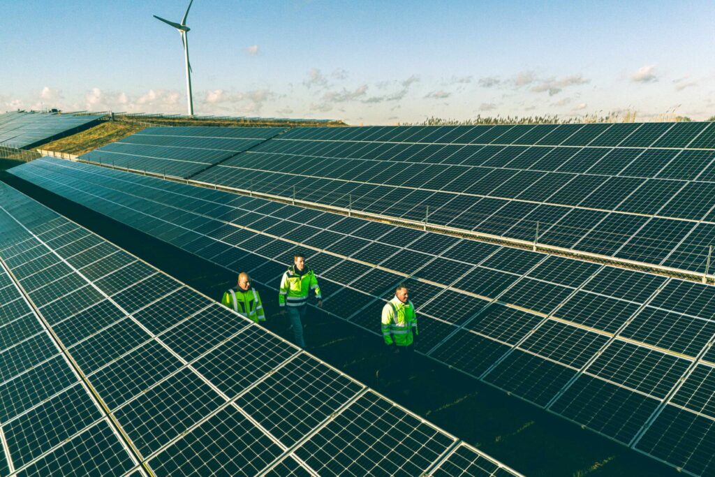 Drie mannen lopen door een zonnepark met een windmolen op de achtergrond. windmolens vlakbij een openbare weg. Dit soort opwekinstallaties zijn te koppelen door middel van cable pooling.